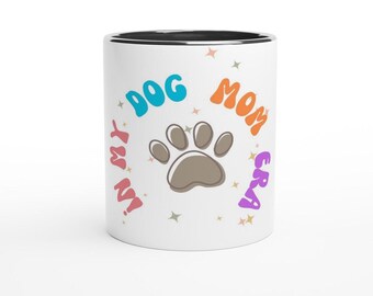 Puede incluir: Taza de cerámica blanca con borde negro. La taza tiene una huella de pata en el centro y el texto "I'm My Dog Mom Era" alrededor de la huella de pata. El texto está en un arcoíris de colores.