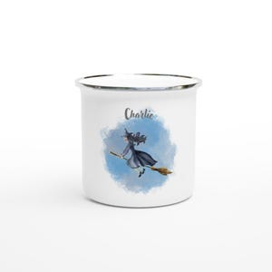 Könnte beinhalten: Weiße Emailtasse mit blauem Aquarellhintergrund und einer Hexe, die auf einem Besen fliegt. Die Tasse hat einen silbernen Rand und der Name "Charlie" ist auf der Vorderseite aufgedruckt.