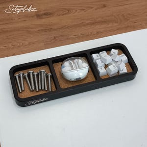 Op de afbeelding: Een zwarte organizerlade met drie compartimenten. Eén sectie bevat zilveren schroeven, een andere een witte oortelefooncase en de derde, witte toetsenbordtoetsen. De lade heeft een kurken basis en de tekst "Setuplabz" in het wit.