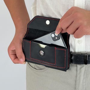 May include: Black leather phone case with red stitching, worn on a belt. The case has a snap closure and a small pocket. A smartphone is being placed inside the case.