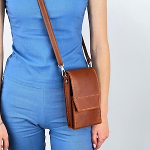 Puede incluir: Un bolso bandolera de cuero marrón con cierre de solapa y correa ajustable. El bolso es rectangular y parece estar hecho de cuero de alta calidad. La correa está sujeta con herrajes de color plateado. El bolso se lleva al hombro.