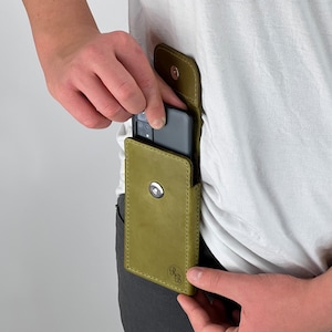 Peut inclure: Étui de téléphone en cuir vert olive avec fermeture à bouton-pression. L'étui est conçu pour être porté à la ceinture. Le téléphone est en cours d'insertion dans l'étui. L'étui a une bordure cousue et un logo dans le coin inférieur droit.