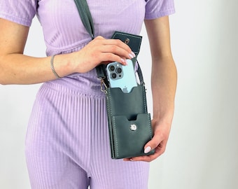 May include: A dark teal leather crossbody phone bag with a matching strap. The bag has a front pocket and a snap closure. A light blue smartphone is visible inside the main compartment. The bag is being held by a person wearing a lavender outfit.