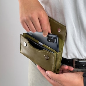 May include: Olive green leather phone case with a magnetic closure. The case is open, revealing a black smartphone. The case is designed to be worn on the belt. The phone case is a practical accessory.