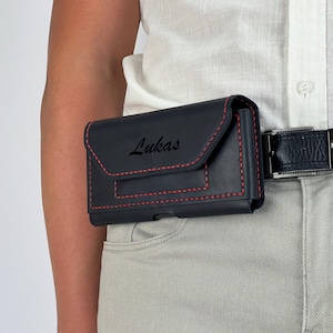 May include: Black leather phone case with red stitching, worn on a belt. The case has a flap closure and the name "Lukas" is printed on the front. The belt is dark blue and the pants are light gray.