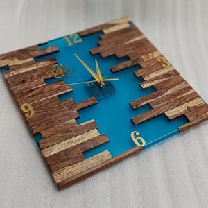 Könnte beinhalten: Quadratische Wanduhr mit blauem Harz in der Mitte und Holzrahmen. Die Uhr hat goldfarbene Ziffern und Zeiger. Der Holzrahmen besteht aus verschiedenen Holzstücken. Das Zifferblatt trägt den Text "Wooden Kings".