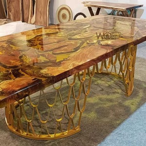 May include: A rectangular wooden table with a clear epoxy resin top. The top features a natural wood grain design with a golden yellow hue. The table has a gold-coloured, decorative metal base.