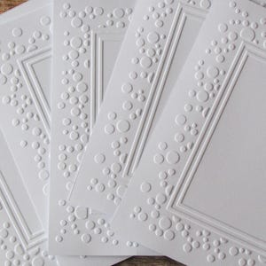 May include: A set of five white square cards featuring an embossed border design. Each card displays a central rectangular frame surrounded by a pattern of raised circles of varying sizes, creating a textured effect on the smooth white paper surface.