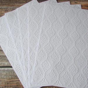 May include: A set of five rectangular white sheets of paper featuring a raised, embossed damask pattern. The paper has a repeating ogee-shaped design with intricate floral details within each section, creating a textured surface across the sheets.