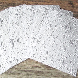 May include: A set of rectangular white paper sheets featuring a raised, embossed floral scrollwork pattern across the entire surface. The paper has a matte finish and a consistent, repeating design of swirling vines and leaves throughout each sheet.