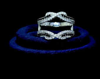 May include: A silver ring setting with a double infinity design. The ring is set with small clear and black stones. The ring is displayed on a blue velvet cushion.