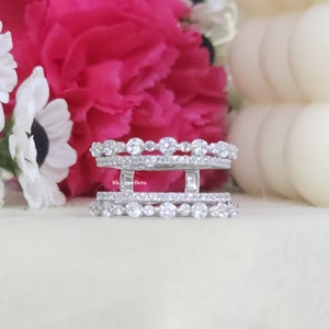 May include: A silver ring with three bands of small, clear stones. The ring is set against a white background with pink flowers.