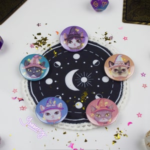 May include: Five round pins with images of cats wearing witch hats. The cats are different colors and have different expressions. The pins are arranged in a circle on a black and white fabric with a crescent moon and stars. The background is white with gold glitter and pink stars.