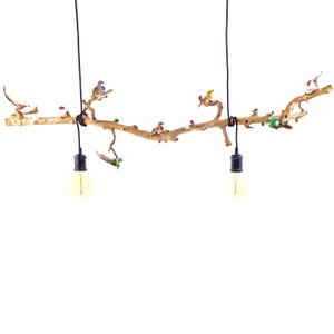 Op de afbeelding: Een unieke lamp met een natuurlijke houten tak versierd met kleurrijke miniatuurvogels. Twee zwarte snoeren hangen de tak op, elk met een gloeilamp. De lamp staat tegen een witte achtergrond, wat de natuurlijke esthetiek benadrukt.