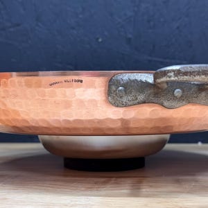 May include: A copper-colored hammered metal bowl with a silver-colored base and a dark handle. The bowl has a hammered texture and is sitting on a wooden surface. The handle is riveted to the bowl. The text "GARANTIE VILLEDFU" is visible on the bowl.