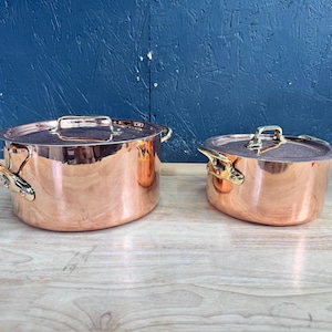 Op de afbeelding: Twee ronde koperen potten met gepolijste oppervlakken en bijpassende deksels. Elke pot is voorzien van twee goudkleurige metalen handgrepen aan de zijkanten. De potten staan naast elkaar en tonen hun metalen afwerking en diepe, ronde constructie.