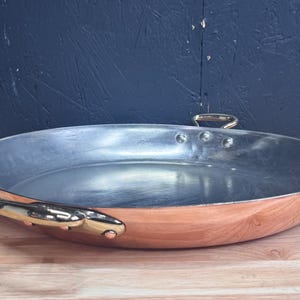 May include: A large, oval copper pan with a silver interior. The pan has two gold-colored handles on the sides and a small handle on the top. The copper exterior has a warm, reddish-brown hue. The pan is sitting on a wooden surface.
