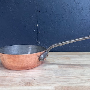 May include: A round copper sauté pan with a hammered exterior finish and a silver-toned metal handle attached with two rivets. The interior of the pan is lined with a metallic silver-colored material, and the number 24 is stamped on the side.