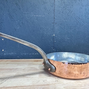 Puede incluir: Una sartén redonda de cobre con un acabado exterior martillado y un mango largo de metal curvado. El interior de la sartén está revestido con una superficie metálica de color plateado. El mango está fijado al lateral con dos remaches.
