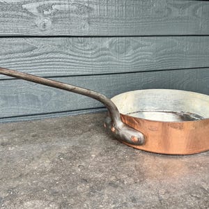 Könnte beinhalten: Eine runde Kupfersautépfanne mit einem langen, dunklen Metallgriff, der mit drei Nieten befestigt ist. Die Außenseite hat ein poliertes Kupferfinish, während die Innenseite mit einem silberfarbenen Metall ausgekleidet ist. Die Pfanne hat einen flachen Boden.