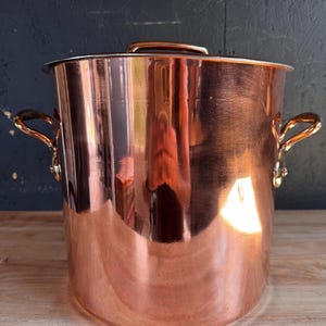Puede incluir: Una olla grande y cilíndrica de cobre con un acabado pulido y reflectante. Cuenta con dos asas metálicas curvas de tono latón fijadas a los lados con remaches y una tapa plana a juego con un asa central en la parte superior.
