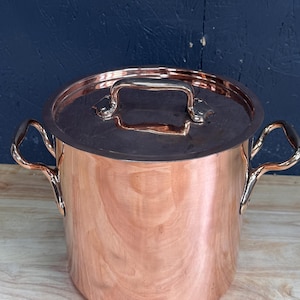 May include: A polished copper stockpot with a matching lid and two side handles. The cylindrical pot features a smooth, reflective metallic surface and a sturdy handle centred on the lid. The design includes riveted handles on both sides.