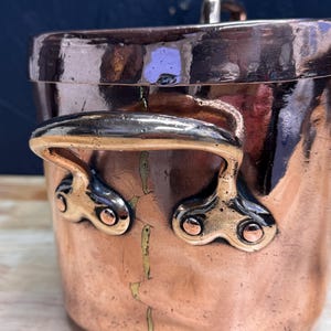 May include: A copper pot with a lid and two handles, reflecting light. The pot's surface is a warm reddish-brown, with gold-coloured handles. A classic piece of kitchenware, the pot is ideal for cooking and adds a touch of vintage charm.