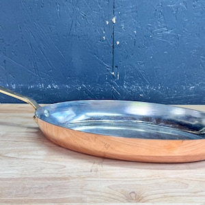 Pode incluir: Uma frigideira oval de cobre com um revestimento interior prateado polido e um cabo longo e fino de latão fixado com um único rebite. O exterior apresenta um acabamento em cobre metálico quente com uma base lisa e arredondada.