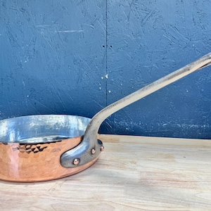 May include: A round copper sauté pan with a hammered exterior finish and a silver-toned metal handle attached with three copper rivets. The interior of the pan is lined with a smooth, metallic silver-colored material, showing a reflective surface.