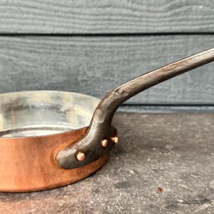 Könnte beinhalten: Eine runde, gehämmerte Kupfersautépfanne mit einer silberfarbenen Innenbeschichtung. Die Pfanne hat einen langen, dunklen, strukturierten Eisengriff, der mit drei sichtbaren Kupfernieten an der Seite befestigt ist und einen Kontrast bildet.