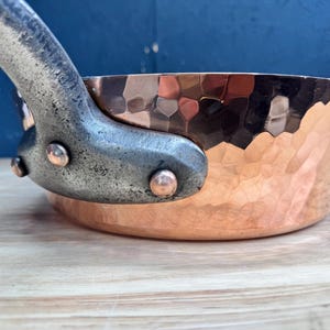 May include: A hammered copper pan with a metallic, textured finish and a curved, weathered-looking handle. The handle is securely attached to the side of the pan with three visible rivets, featuring a distinct contrast between the shiny copper body and the dark, matte, industrial-style handle.