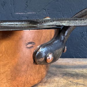 Puede incluir: Primer plano de una cacerola de cobre con un asa y una tapa de metal oscuro. La olla tiene un exterior de cobre brillante y un asa de metal oscuro sujeta con remaches de cobre. La tapa también es de metal oscuro. La olla tiene un sello que dice "ARTEVELDE ANTWERPEN".