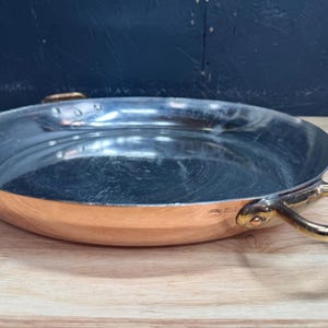 Peut inclure: Une poêle ronde et peu profonde en cuivre avec un revêtement intérieur argenté poli. L'extérieur présente une finition en cuivre métallique chaud. Deux poignées en métal doré sont fixées aux côtés opposés avec des rivets visibles.
