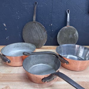 Op de afbeelding: Een verzameling antieke koperen kookgerei, waaronder twee ronde pannen met lange handvatten en drie koperen potten met ijzeren handvatten. De koperen potten hebben een glanzend interieur en een roodbruin exterieur. De pannen en potten zijn gerangschikt op een houten oppervlak.