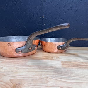 Op de afbeelding: Twee koperen steelpannen met gehamerde buitenkanten en geklonken ijzeren handvatten. De pannen hebben een heldere koperkleur en staan op een houten oppervlak. De handvatten zijn donkerbruin en gebogen.