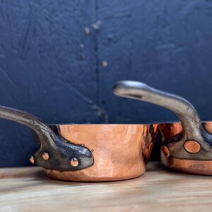 Op de afbeelding: Twee koperen steelpannen met gehamerde buitenkanten en donkere metalen handvatten. De pannen hebben een klassiek ontwerp, met klinknagels die de handvatten aan de koperen lichamen bevestigen. De handvatten buigen omhoog. Ze staan op een houten oppervlak.
