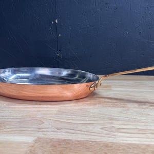 Könnte beinhalten: Eine kupferfarbene Bratpfanne mit einem langen, goldfarbenen Griff. Der Innenraum ist silberfarben. Die Pfanne steht auf einer Holzoberfläche mit dunklem blauem Hintergrund. Küchenutensilien.