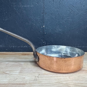Op de afbeelding: Een ronde koperen sauteerpan met een gehamerde buitenkant en een zilverkleurige binnenkant. De pan is voorzien van een lange, gebogen metalen handgreep die met een klinknagel aan de zijkant is bevestigd voor een stevige grip.
