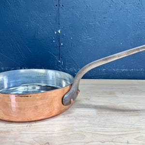 May include: A round copper sauté pan with a hammered exterior finish and a silver-toned interior lining. The pan features a long, curved metal handle attached to the side with two visible rivets, providing a sturdy grip for cooking tasks.