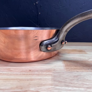 Op de afbeelding: Een koperen steelpan met een geklonken, donker metalen handvat. De pan heeft een glanzend, reflecterend oppervlak en een donker interieur. De handgreep buigt omhoog en is met koperen klinknagels aan de pan bevestigd.