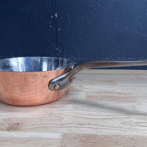 Puede incluir: Una sartén de cobre con interior plateado pulido y un mango de metal largo y curvado sujeto con dos remaches de cobre. El exterior de la sartén presenta una pequeña marca estampada que dice Made in France.