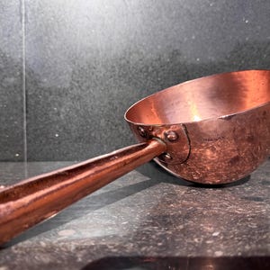 Puede incluir: Un cucharón de cobre pulido con un cuenco redondeado y un mango largo y cónico. La superficie metálica presenta una sutil textura martillada en toda su extensión. Una pequeña marca circular del fabricante está estampada en el cuenco.