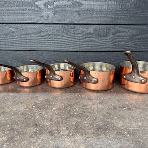 May include: A set of five graduated copper saucepans with hammered exterior finishes, silver-toned interior linings, and dark metal handles secured by rivets. Each pan is stamped with the letters HA on the side near the handle attachment point.