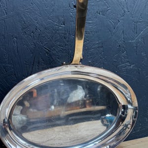 May include: An oval-shaped copper pan with a polished silver-toned interior and a long, slender handle extending from the top. The pan features a reflective surface and a sturdy metal construction with a visible rivet connecting the handle to the body.