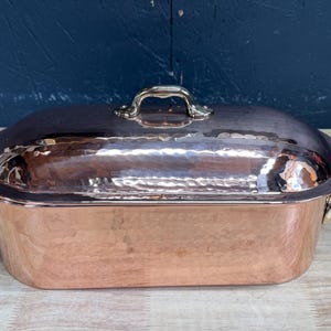 May include: An oval-shaped hammered copper roasting pan with a matching lid. The pan features two brass side handles and a central brass handle on the lid. The exterior surface has a polished metallic finish with visible hammered indentations.