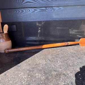 May include: A long, smooth wooden stirring paddle with a rounded handle end and a flat, rectangular blade. Beside it sits a two-toned brown ceramic crock containing two wooden kitchen utensils with slotted heads, all featuring a natural wood grain.