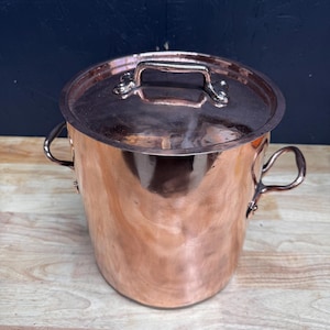 Op de afbeelding: Een koperen kookpot met een deksel. De pot heeft twee zijhandvatten en een handvat op het deksel. Het koper heeft een glanzend, reflecterend oppervlak. De pot staat op een houten oppervlak.