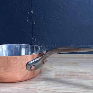 Pode incluir: Uma frigideira sauté redonda de cobre com interior metálico polido e um cabo de metal longo e resistente fixado com três rebites visíveis. A superfície exterior é de cobre liso e um pequeno selo é visível perto da fixação do cabo.