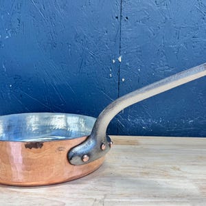 Puede incluir: Una sartén redonda de cobre con un interior metálico pulido y un mango largo y curvado de metal plateado unido con tres remaches visibles. El exterior de la sartén presenta un acabado de cobre marrón rojizo cálido con un pequeño logotipo.