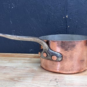 May include: A copper saucepan with a silver handle attached with copper rivets. The pot has a slightly rounded shape and a smooth, polished surface. The letters "GE" are stamped on the side of the pot. The background is a dark blue wall and a wooden surface.